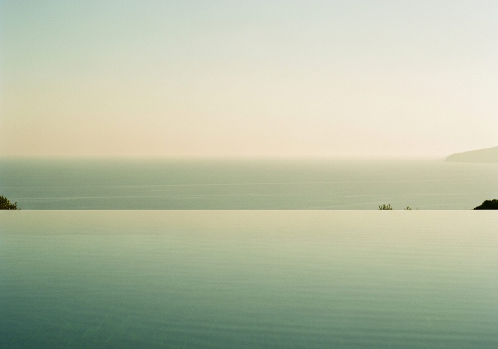 A still infinity pool at dawn facing a hazy sage-green coastline